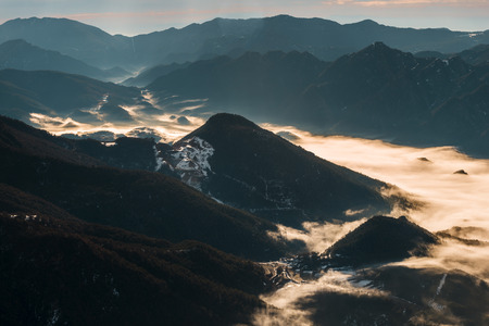 Back-lit view of a valley on a winter morning. The cold weather makes the fog stay between the mountains covering the valley like a sea of fogの写真素材