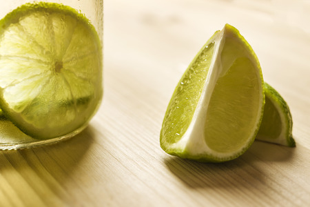horizontal photo of some slices of lime next to a glass jar with lemonade, this one is illuminated by sunlight and on a wooden tableの写真素材
