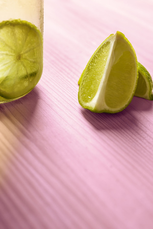 vertical photo with copy space of some slices of lime next to a glass jar with lemonade, this one is illuminated by sunlight and on a pink wooden tableの写真素材