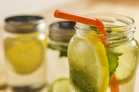 close-up of the top of a glass jar with ice water, lemon, lime, mint leaves and a cane to drink, at the bottom and out of focus are other lemonadesの写真素材