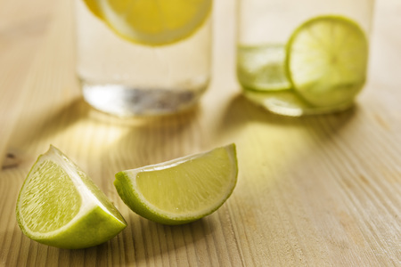 two slices of lemon on a wooden table with homemade citrus sodas unfocused in the background and illuminated by sunlight, the glasses are filled with water with slices of lemon and limeの写真素材