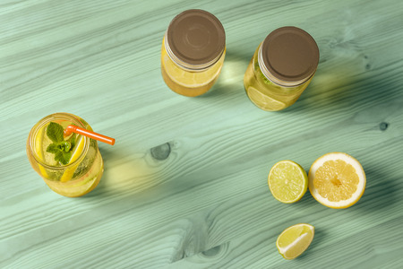 Flat Lay of lemonade with mint and a cane to drink in a glass jar illuminated by sunlight, is on a turquoise wooden table with two other jars with lid, some pieces of lemon and lime and copy spaceの写真素材