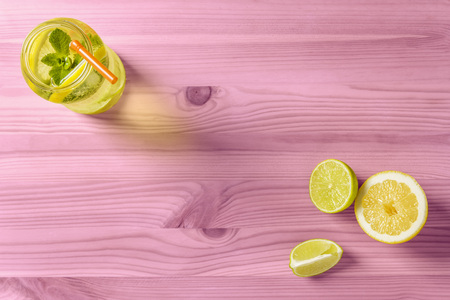 flat lay of homemade lemonade with mint and a cane to drink in a glass jar lit by sunlight, it's on a pink wooden table, there are also a few pieces of lime and lemon and copy spaceの写真素材