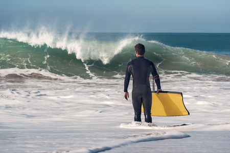 Man on the seashore prepares to surf, wears a black neoprene wetsuit and in his hand wears a yellow bodyboard. the water covers him to the knee while watching a big wave breakingの写真素材