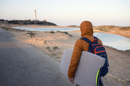 Surfer with his surfboard walks towards the sea sheltered with a brown coat, it is winter and it is...