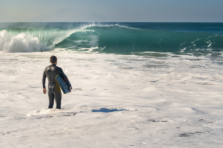 Man on the seashore prepares to surf, wears a black neoprene suit and in his hand has a blue bodyboard. the water covers him to the knee while watching a huge wave breakingの写真素材