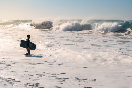 Man on the seashore prepares to surf, wears a black neoprene suit and has a blue bodyboard. the...
