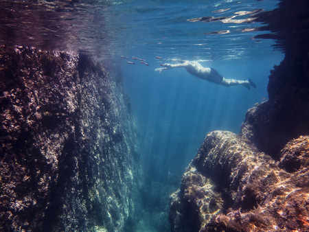 Underwater view of a man swimming placidly in the sea with some fish. Sun rays are drawn in the crystal clear water and illuminate the rocks and curly surface of the blue oceanの写真素材