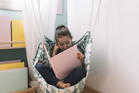 little girl laughing while reading a book near the window, funny lovely child having fun in her kids room, copy space for textの写真素材