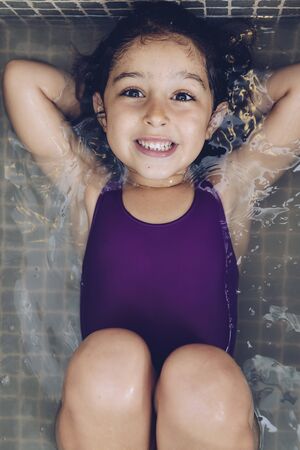 vertical photo of a beautiful little girl having fun and smiling lying in the tub while taking a bath in the bathtub, kids hygiene conceptの写真素材