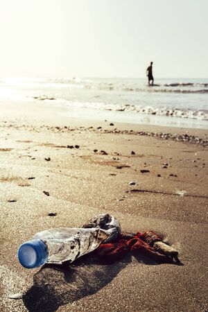 vertical photo of a plastic bottle and garbage lying on the shore of the beach polluting the sea and...