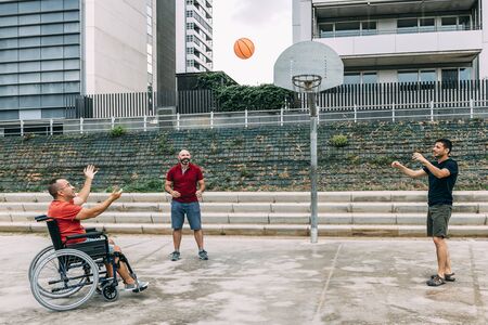 joyful disabled man in wheelchair playing basketball with friends with a ball, concept of adaptive...