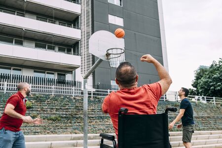 joyful disabled man in wheelchair throwing the ball to basket with two friends, concept of adaptive sports and physical activity, rehabilitation for people with physical disabilitiesの写真素材