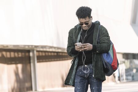 black young man with earphones walking by the city and texting with a mobile phone, technology and...
