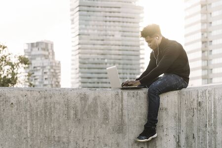 handsome black man with white earphones working typing in a computer sitting outdoors in the city at sunset, lifestyle and technology concept using internet electronic deviceの写真素材
