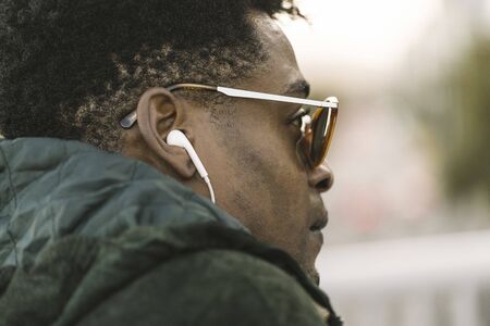 attractive young black african man with green jacket and sunglasses listening music with white earphones, city lifestyle conceptの写真素材