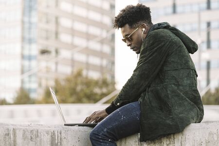 handsome young student with sunglasses and earphones working on laptop sitting outdoors in the city, lifestyle and technology concept using internet electronic deviceの写真素材
