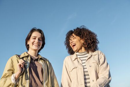 two young women laughing animatedly against a background of blue sky, concept of female friendship...