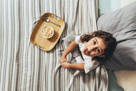 top view of a teasing little girl sticking out her tongue with a sunday breakfast in her parents' bed. Kid enjoying a bowl of cereal at home, concept of healthy living and foodの写真素材