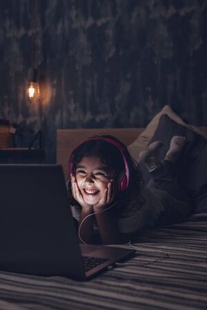 vertical photo of a happy little girl lying in bed smiling while watching a movie on a computer in the darkness, has a pink headset, child and technology concept, copy space for textの写真素材