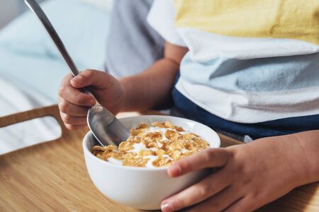 detail of a little girl sitting on the bed holding a spoon and a bowl with cereal and milk for breakfast. Kid enjoying with healthful food, healthy lifestyle conceptの写真素材