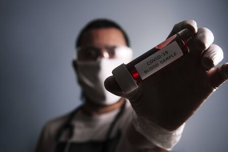 portrait of a man with a protective medical mask showing a medical test tube with blood sample infected with covid-19, concept of coronavirus research, selective focus on vialの写真素材
