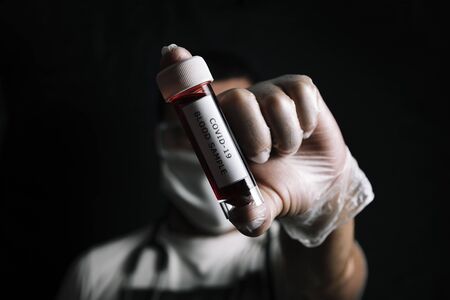dark photo of a doctor with protective gloves and medical mask showing a vial with blood sample infected with covid-19, concept of coronavirus research, selective focus on vialの写真素材