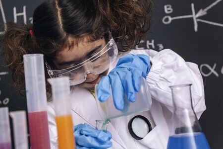 little scientist girl with goggles and gloves in lab coat mixing chemical liquids in flasks, blackboard background with science formulas, explosion in the laboratory, back to school conceptの写真素材