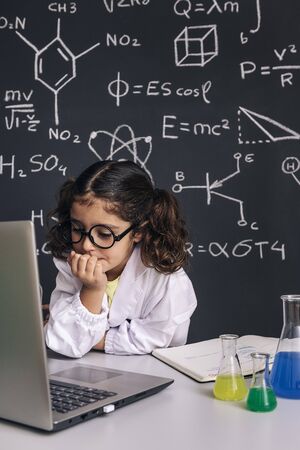 little scientist girl with glasses in lab coat looking to the computer, on school blackboard...