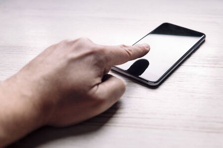 hand with the finger touching the screen of a smart phone with blank screen on white wooden table,...