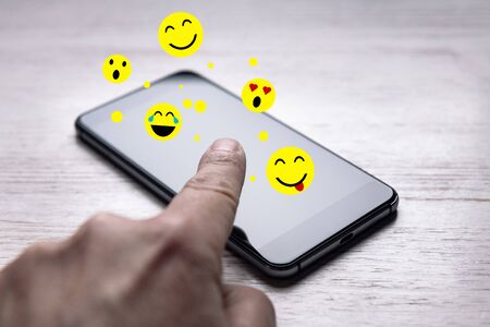 finger touching the screen of a phone that emoticons come out of on white wooden table, social media...