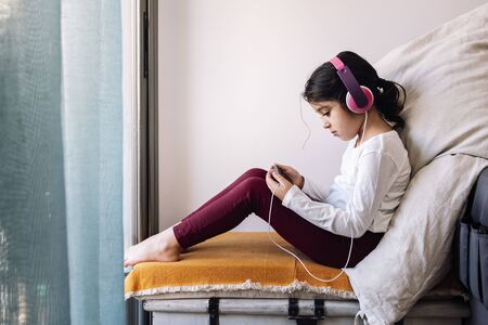 little girl sitting in front of her home window with headphones watching videos on the phone, home entertainment for children conceptの写真素材