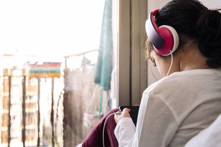 little girl sitting in front of her house balcony with headset watching videos on the phone, home entertainment for children concept, copy space for textの写真素材