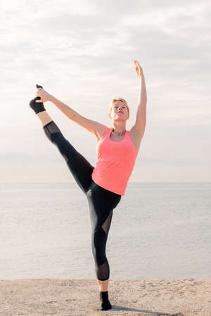 vertical photo of a beautiful blonde woman practicing balancing and yoga stretches at sunrise in...