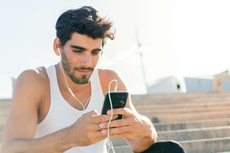 handsome sporty man with earphones selects music on his phone to listen during his workout, urban sport concept and healthy lifestyle, copy space for textの写真素材