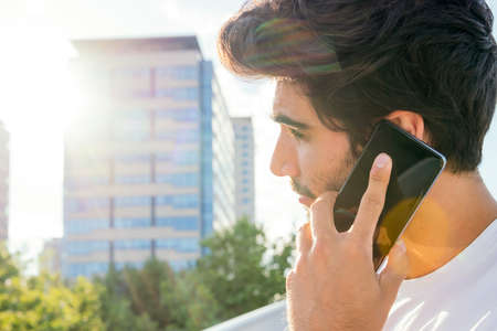 handsome young man talking on his smartphone in front of city buildings at sunset, technology...