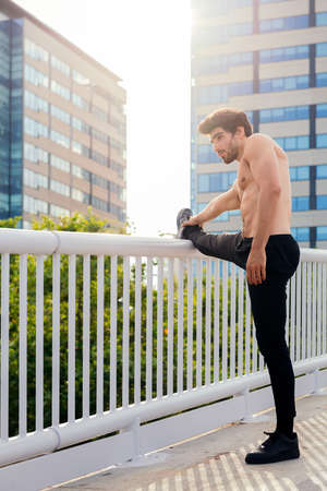 vertical photo of a handsome shirtless sportsman stretching his leg muscles in front of city buildings, concept of urban sport and healthy lifestyle, copy space for textの写真素材