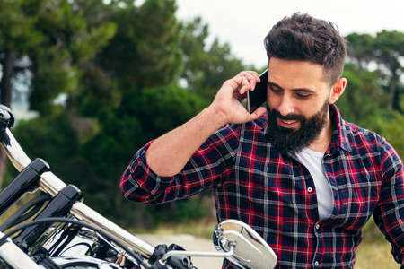 portrait of a handsome bearded man talking on his mobile phone sitting on his motorcycle, concept of freedom and biker lifestyle, copy space for textの写真素材