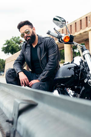 vertical photo of a man in leather jacket rests by the side of the road sitting on his motorcycle, concept of freedom and biker lifestyleの写真素材