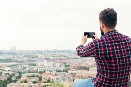 back view of a man taking a photo with his mobile phone of the city views from a rooftop, concept of...