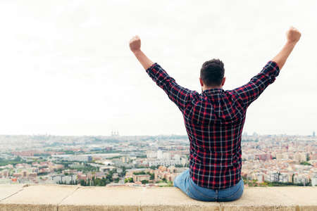 rear view of a man sitting on the rooftop observing the city with arms up, concept of freedom and disconnection, copy space for textの写真素材