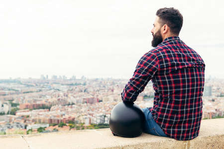 handsome bearded man sitting on the rooftop with a motorcycle helmet looking the city at his feet, concept of freedom and disconnection, copy space for textの写真素材