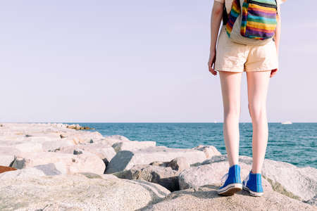 unrecognizable young traveler girl with sneakers and backpack walking on the rocks by the sea,...