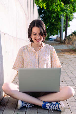 vertical photo of a young woman with earphones working with a laptop sitting next to a gray wall, technology concept and blogging lifestyleの写真素材