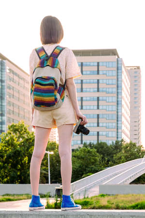 rear view of a young traveler woman with backpack and camera standing in front of the city, concept of youth and digital nomad lifestyle, vertical photoの写真素材