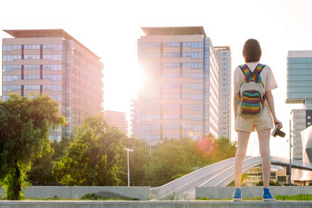 rear view of a young traveler woman with backpack and camera standing in front of the city, concept of youth and digital nomad lifestyle, copy space for textの写真素材
