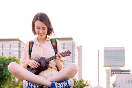 beautiful young woman in sneakers playing ukulele at sunset in the city, amateur music concept and...