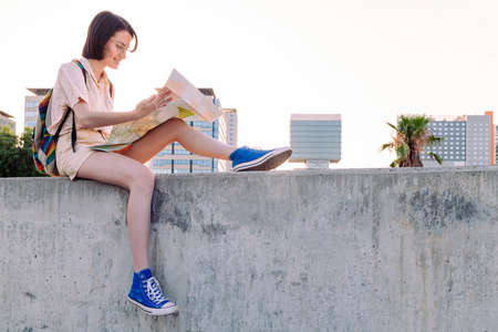 happy young woman with backpack looking a map sitting in front of the city, concept of youth and nomad lifestyle, copy space for textの写真素材