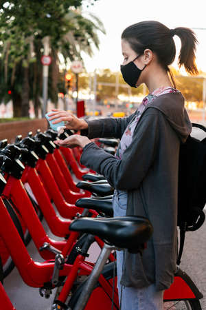 vertical photo of a young girl with protective mask cleaning her hands with disinfectant gel next to an urban bicycle rental parking, concept of protection against covid and sustainable mobilityの写真素材