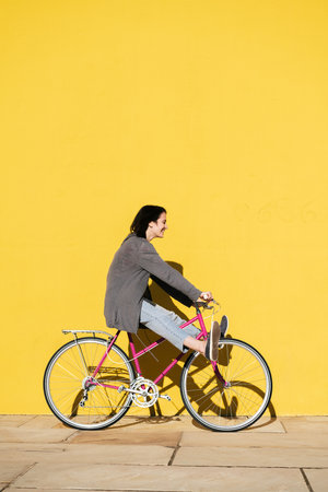 vertical photo of a happy young girl smiling as she plays on her retro pink bike next to a colorful yellow wall, concept of active lifestyle and sustainable mobility, copy space for textの写真素材